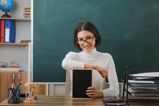 教室で学校のツールをオンにして机に座ってミニ黒板を保持している眼鏡をかけて笑顔の若い女性教師