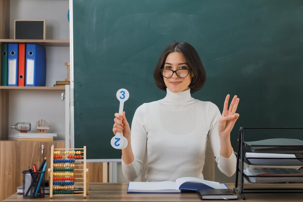 smiling young female teacher wearing glasses holding number fan sitting at desk with school tools on in classroom