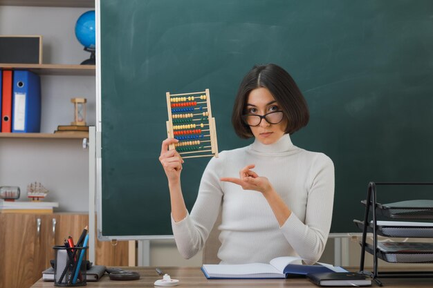 smiling young female teacher wearing glasses holding and points at abacus sitting at desk with school tools on in classroom