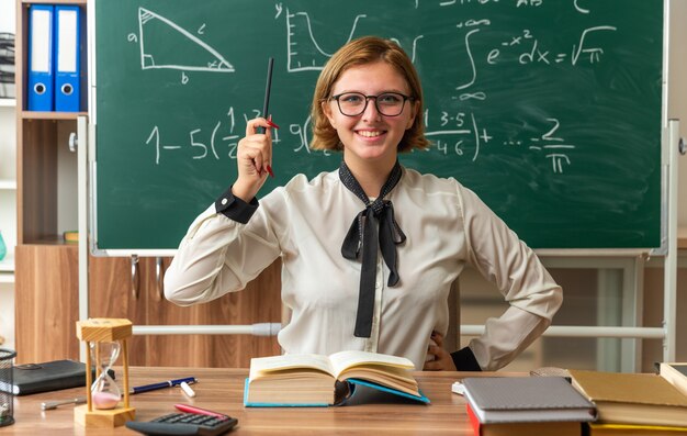 smiling young female teacher wearing glasses sits at table with school supplies holding pencil putting hand on hip in classroom