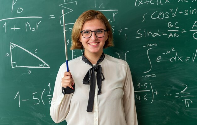 Smiling young female teacher wearing glasses standing in front blackboard holding pointer stick in classroom