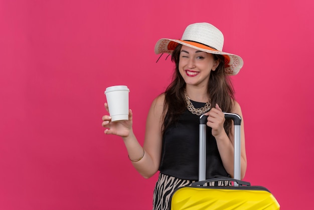 Улыбающаяся молодая путешественница в черной майке в шляпе мигает и держит чашку кофе на красной стене