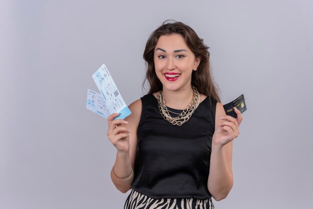 白い壁にチケットとクレジットカードを保持している黒いアンダーシャツを着て笑顔の若い女性旅行者