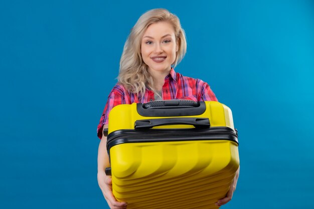 Smiling young female traveler wearing red shirt holding suitcase on isolated blue wall