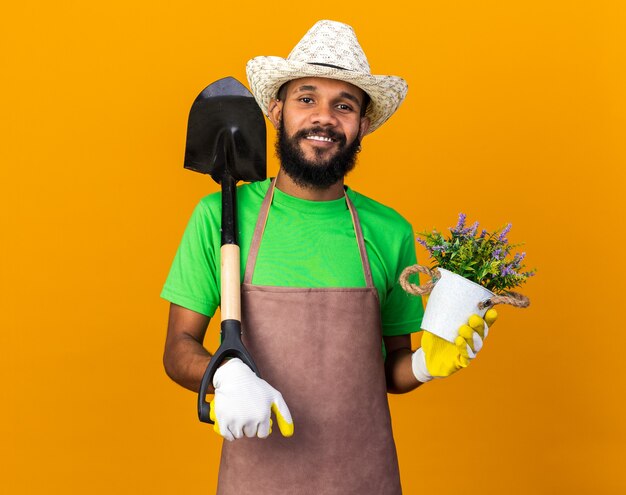 Smiling young gardener afro-american guy wearing gardening hat and gloves holding spade with flower in flowerpot 