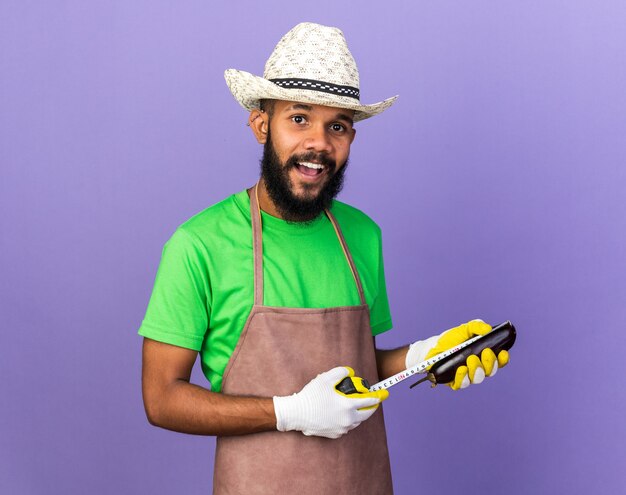 푸른 벽에 격리된 줄자로 가지를 측정하는 원예용 모자와 장갑을 끼고 웃고 있는 젊은 정원사 아프리카계 미국인 남자