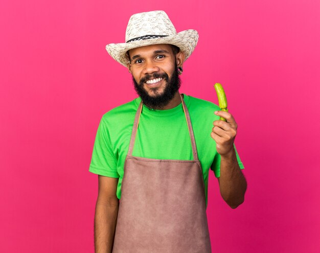 Smiling young gardener afro-american guy wearing gardening hat holding broke pepper isolated on pink wall