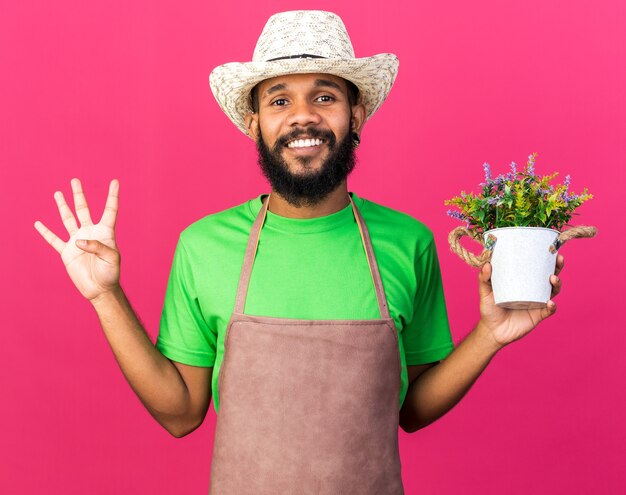 Smiling young gardener afro-american guy wearing gardening hat holding flower in flower pot showing four isolated on pink wall