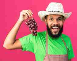 Free photo smiling young gardener afro-american guy wearing gardening hat holding grappes isolated on pink wall