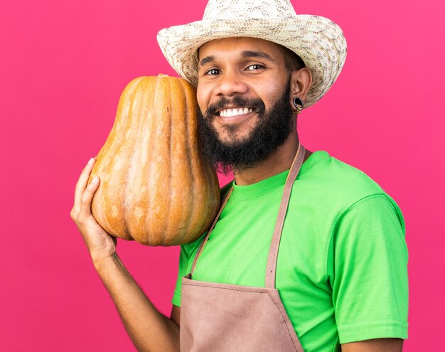 カボチャを保持しているガーデニングの帽子をかぶっている若い庭師のアフリカ系アメリカ人の男を笑顔