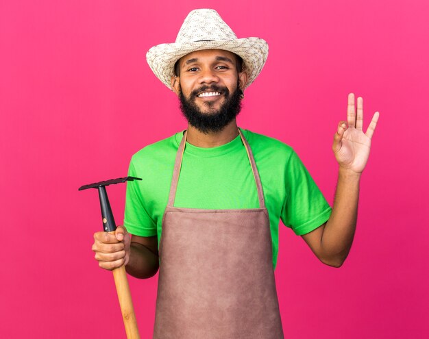 Smiling young gardener afro-american guy wearing gardening hat holding rake showing okay gesture isolated on pink wall