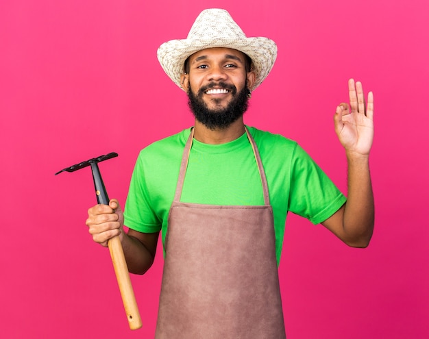 Sorridente giovane giardiniere afroamericano che indossa un cappello da giardinaggio che tiene in mano un rastrello che mostra un gesto ok isolato su una parete rosa