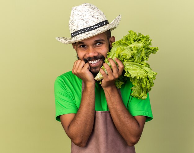Smiling young gardener afro-american guy wearing gardening hat holding salad isolated on olive green wall