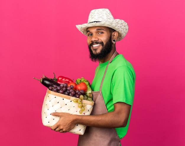 Smiling young gardener afro-american guy wearing gardening hat holding vegetable basket isolated on pink wall