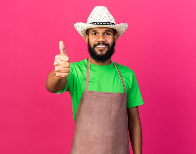 Улыбающийся молодой афро-американский парень садовник в садовой шляпе показывает большой палец вверх изолирован на розовой стене