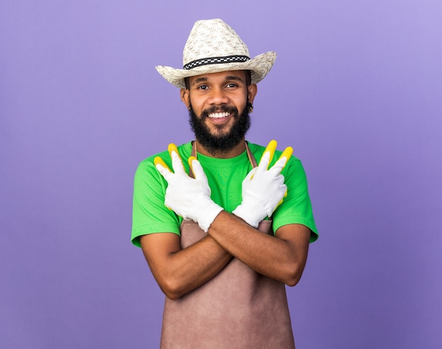 Smiling young gardener afro-american guy wearing gardening hat with gloves showing peace gesture isolated on blue wall