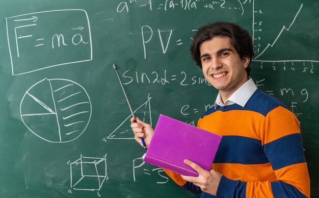 smiling young geometry teacher standing in front of chalkboard in classroom holding book pointing with pointer stick at chalkboard looking at front