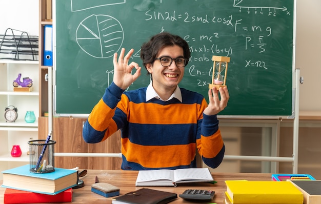 Foto gratuita sorridente giovane insegnante di geometria con gli occhiali seduto alla scrivania con materiale scolastico in aula tenendo la clessidra guardando davanti facendo segno ok