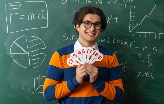smiling young geometry teacher wearing glasses standing in front of chalkboard in classroom holding number fans looking at front