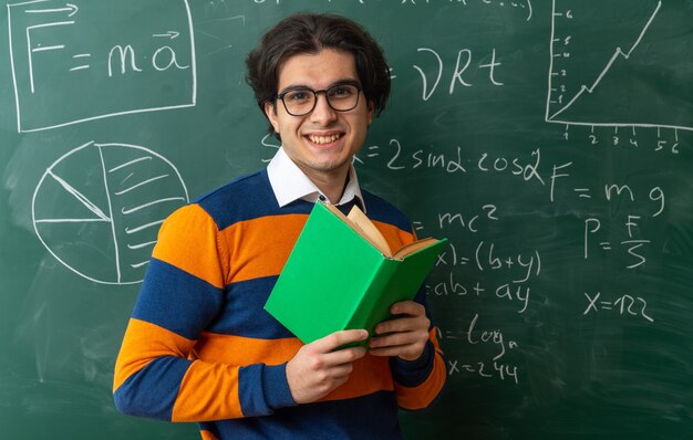 smiling young geometry teacher wearing glasses standing in front of chalkboard in classroom holding open book looking at front