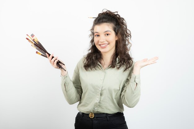 Smiling young girl artist showing opened palm and holding a bunch of paintbrushes . 