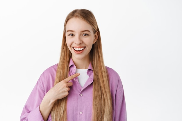 Smiling young girl candidate, pointing at herself and looking confident, self-promoting, talking personal achievements, bragging, standing over white wall