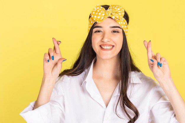Smiling young girl crossed her fingers and standing on yellow background