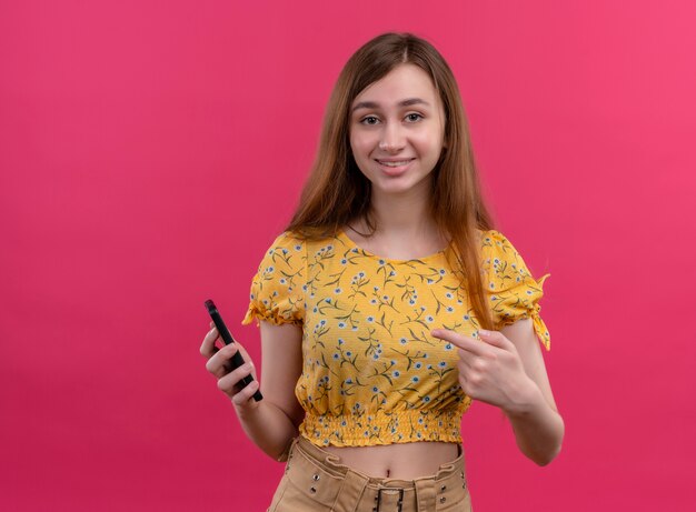 Улыбающаяся молодая девушка держит мобильный телефон и указывает на него на изолированной розовой стене с копией пространства