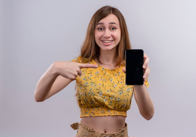 Smiling young girl holding mobile phone and pointing at it on isolated white wall