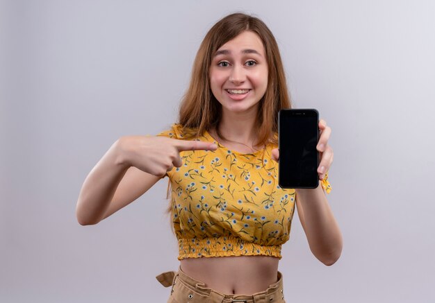Улыбающаяся молодая девушка держит мобильный телефон и указывает на него на изолированной белой стене