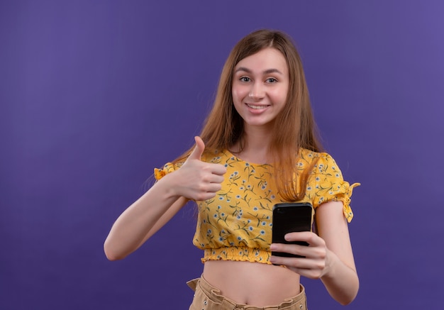 Smiling young girl holding mobile phone and showing thumb up on isolated purple wall