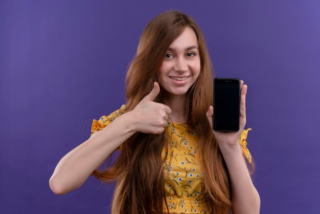 휴대 전화를 들고 고립 된 보라색 벽에 엄지 손가락을 보여주는 어린 소녀 미소