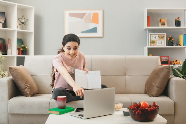 Smiling young girl holding notebook sitting on sofa behind coffee table looking and points at laptop in living room