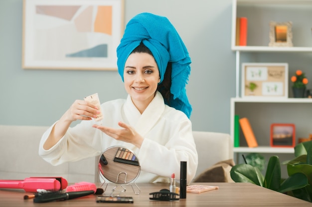 Sorridente ragazza con crema tonificante capelli avvolti in un asciugamano seduto a tavola con strumenti per il trucco in soggiorno