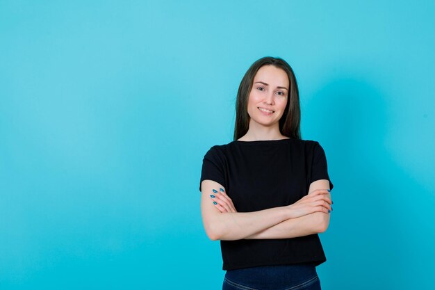 Smiling young girl is looking at camera by crossing hands on blue background