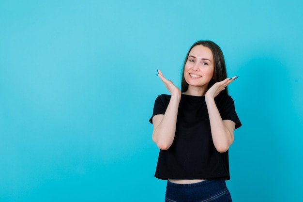 Smiling young girl is looking at camera by holding hands around face on blue background