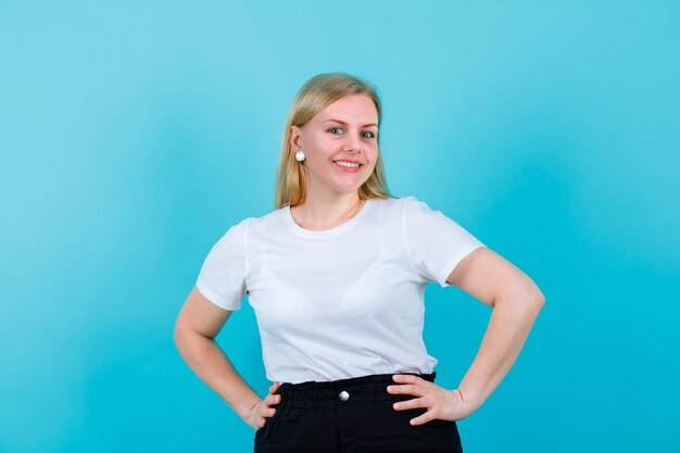 Smiling young girl is looking at camera by putting hands on waist on blue background