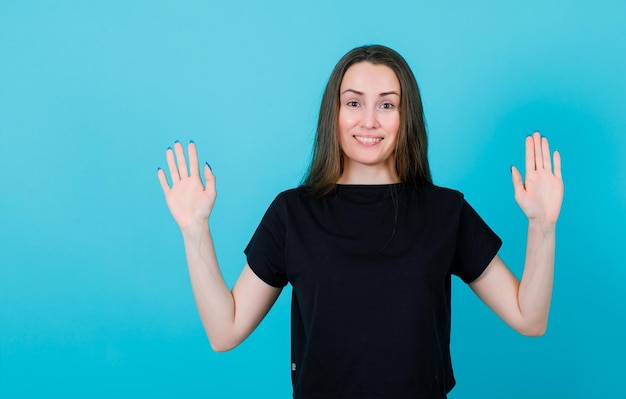 Smiling young girl is looking at camera by rasing up hands on blue background