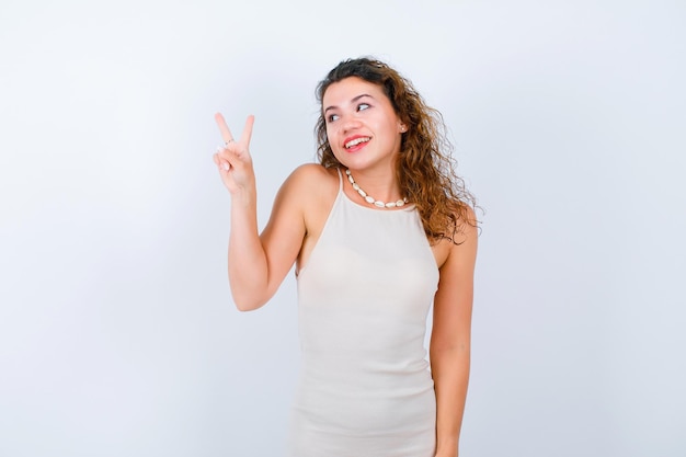 Smiling young girl is looking left by showing victory gestures on white background