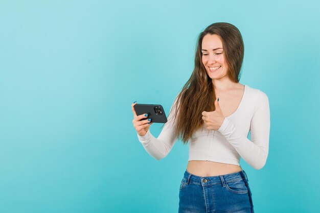 Smiling young girl is looking at smartphone screen by showing perfect gesture on blue background