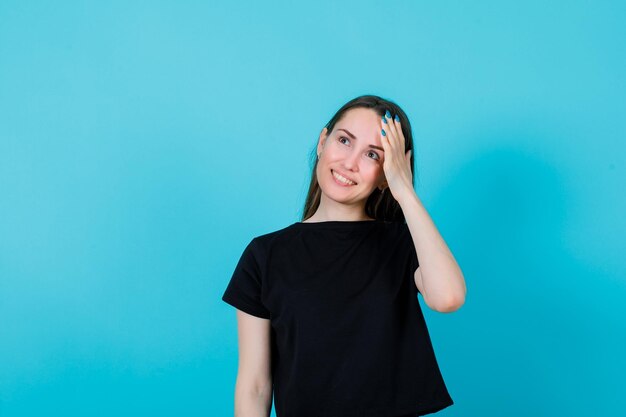 Smiling young girl is looking up by putting hand on head on blue background