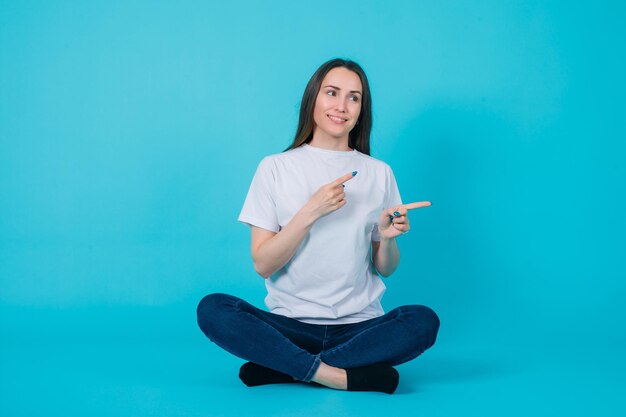 Smiling young girl is pointing right with forefingers by sitting on floor on blue background