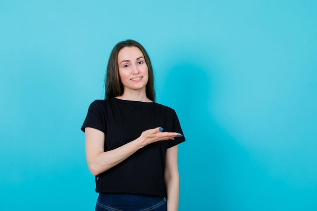 Smiling young girl is pointing right with hand on blue background