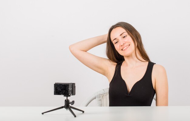 Smiling young girl is posing at little camera by holding hand behind head on white background