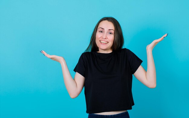Smiling young girl is raising up hands by opening wide them on blue background