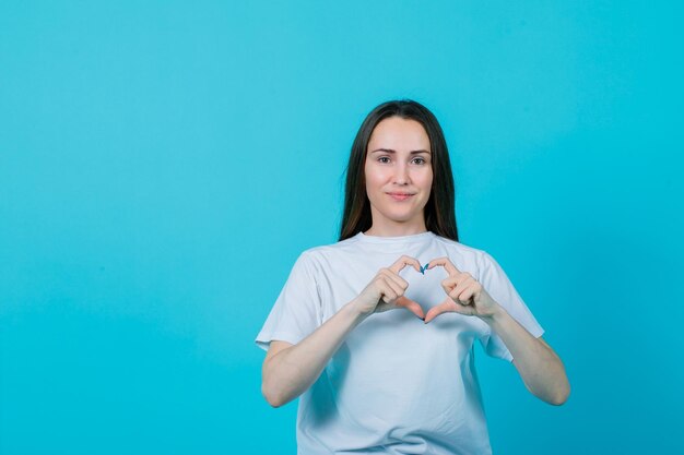 Smiling young girl is showing heart gesture on blue background