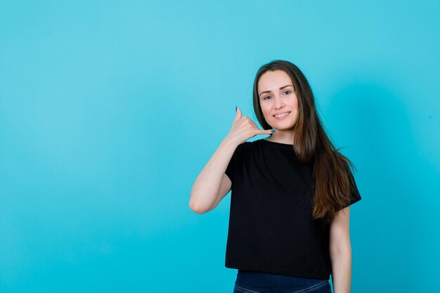 Smiling young girl is showing phone gesture by holding hand near ear on blue background