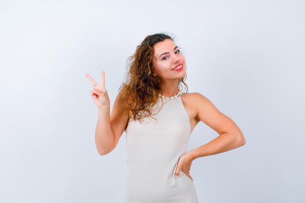 Smiling young girl is showing two gesture and putting other hand on waist on white background