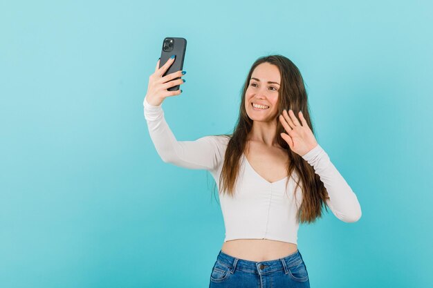 Smiling young girl is taking selfie by showing hi gesture on blue background