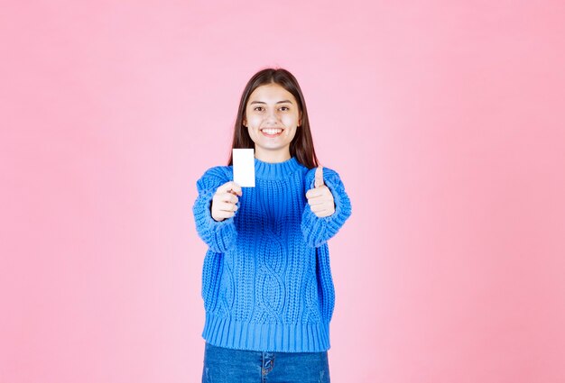 분홍색 벽에 엄지 손가락을 보여주는 카드와 함께 웃는 어린 소녀 모델.